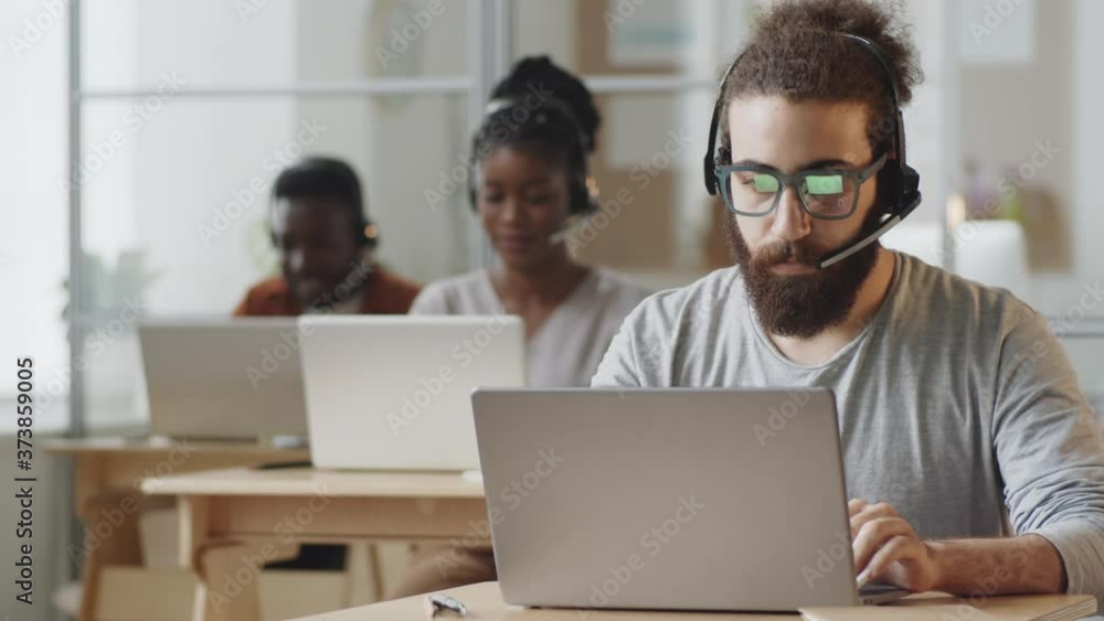 Young bearded man in headset typing on laptop, then posing for camera and smiling while working at desk in customer service center Young bearded man in headset typing on laptop, then posing for camera and smiling while working at desk in customer service center