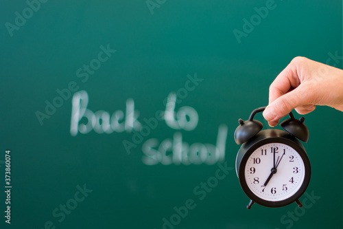 On the palm of your hand, against the background of a blackboard with the inscription back to school, soft focus, there is a black alarm clock. Back to school concept.