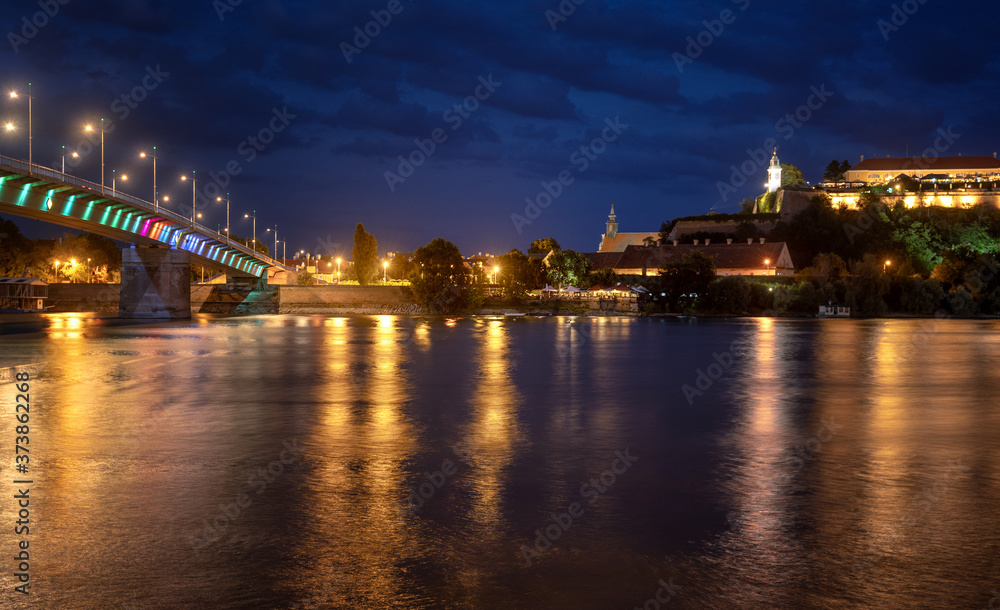 Petrovaradin fortress in Novi Sad, Serbia illuminated with colorful street lights and reflection in the Danube river and rainbow bridge water