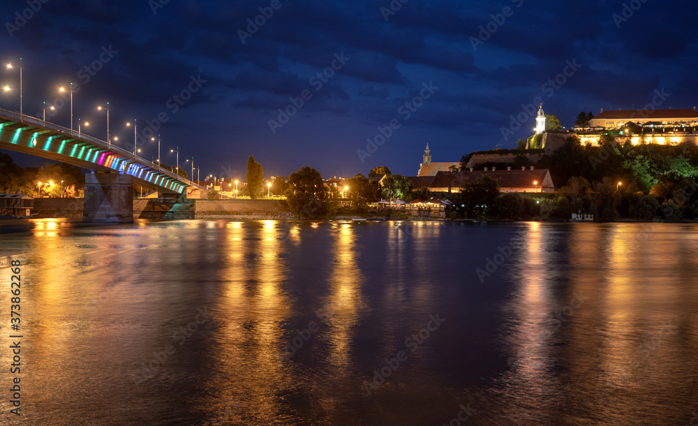 Obraz premium Petrovaradin fortress in Novi Sad, Serbia illuminated with colorful street lights and reflection in the Danube river and rainbow bridge water
