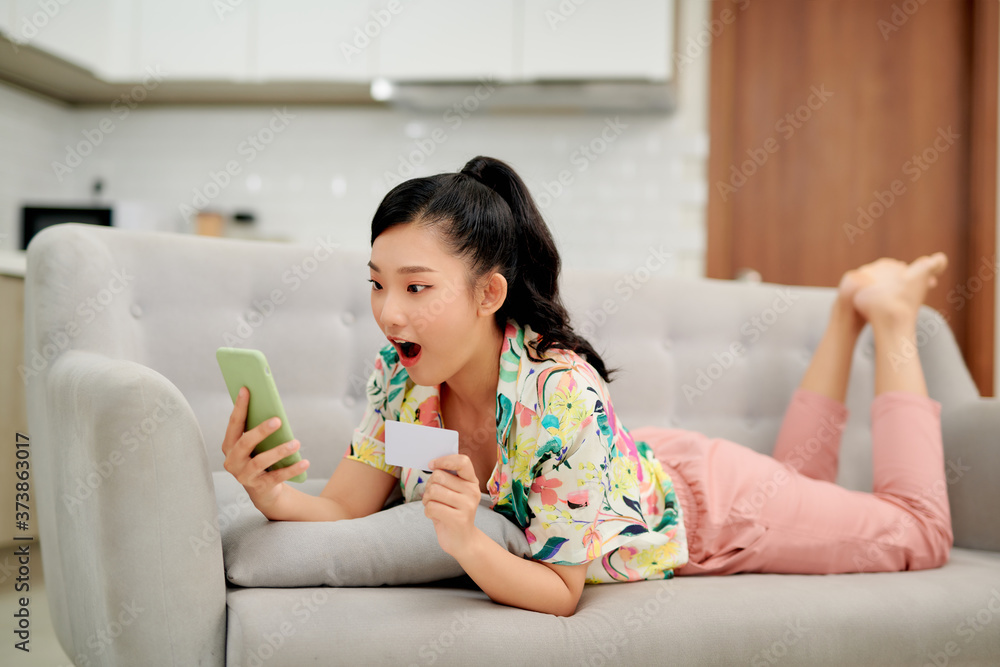 Image of optimistic excited young woman indoors at home using mobile phone holding credit card on sofa. Image of optimistic excited young woman indoors at home using mobile phone holding credit card on sofa.