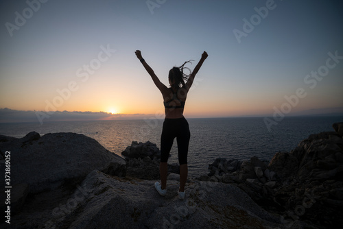 An young active sporty athlete woman with firm fit body is exulting of happiness satisfied with her achievement after running and jogging workout on a top of rock with seascape at sunset.