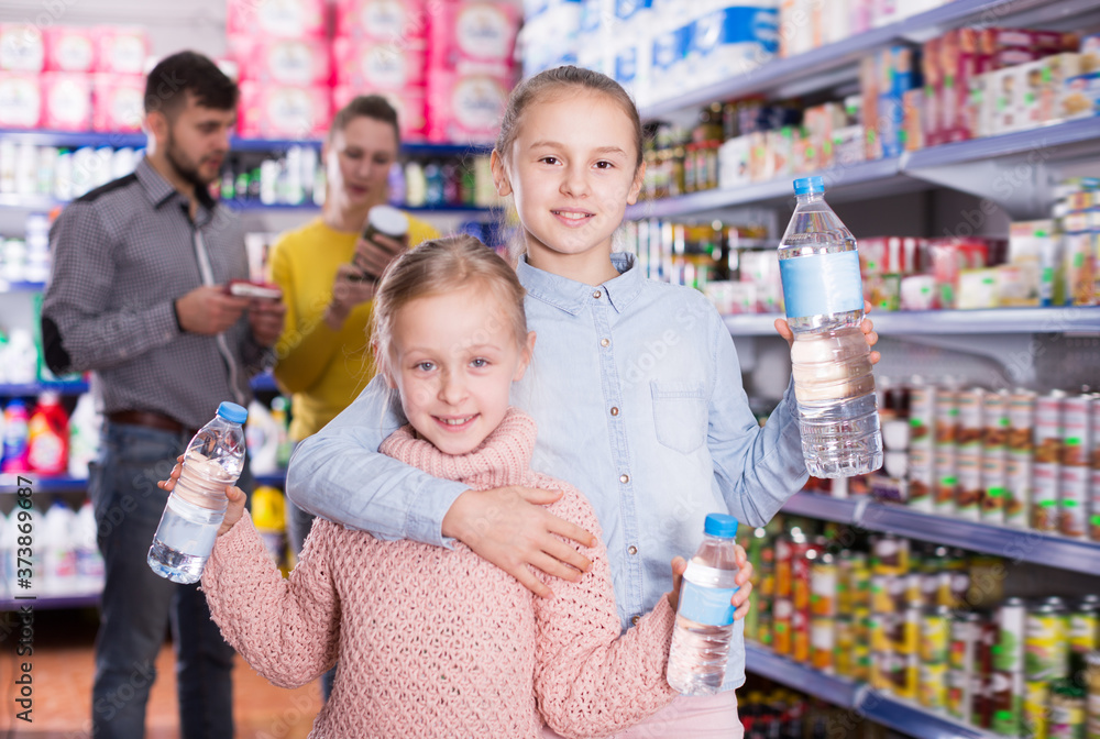 Two small glad sisters holding bottled water during family shopping in supermarket