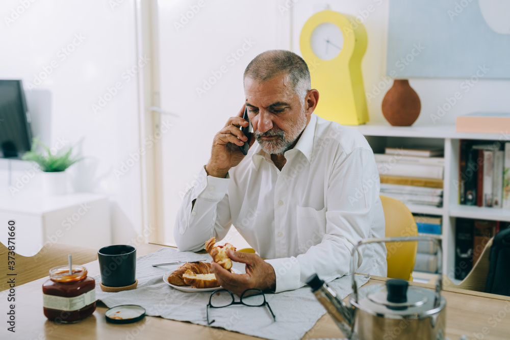 middle aged man talking on mobile phone while having breakfast at home middle aged man talking on mobile phone while having breakfast at home