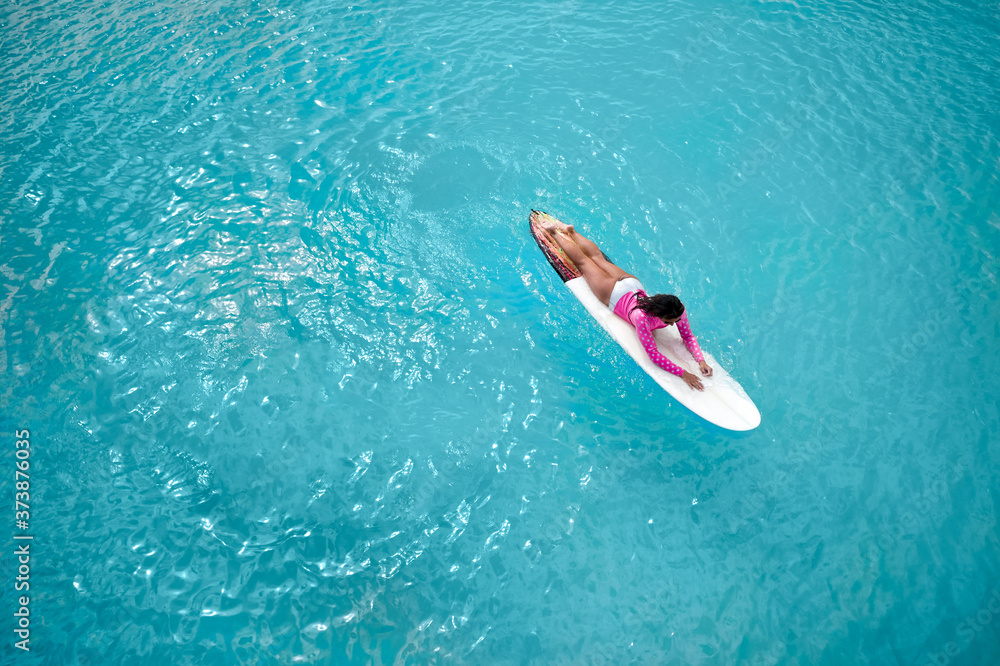 Brunette surfer girl on a long surfboard relax in the blue transparent ocean. From above aerial drone shot.