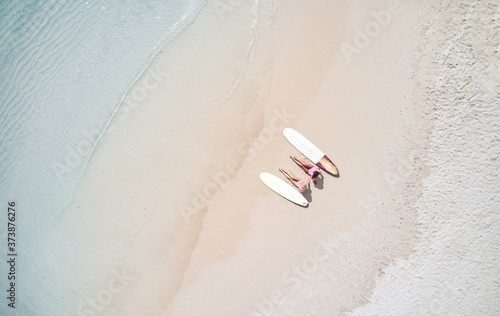 Blonde and brunette surfer girls on a long surfboards relax on the empty beach near the blue transparent ocean. From above aerial drone shot.