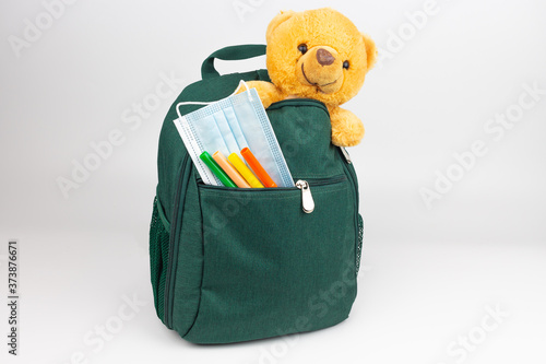 Green school backpack with a teddy bear and in markers and protective mask in the front pocket

