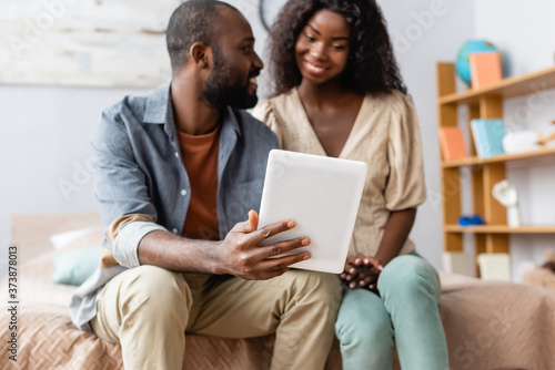 selective focus of african american man showing digital tablet to wife while sitting on bed