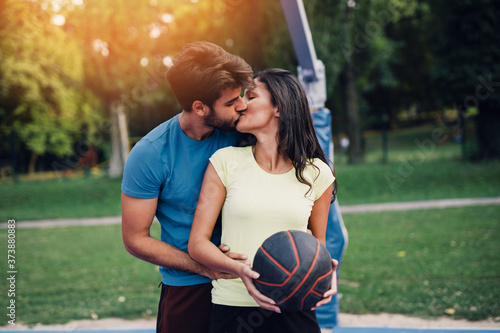 Beautiful young couple kissing and enjoying together and playing on basketball court. Bright sunny summer day.