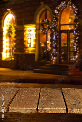 Old shabby board on a background of blurred Christmas market. Brown table top in sharpness. Christmas decorations, lights and lanterns in the background. Layout for design. Free space for products.