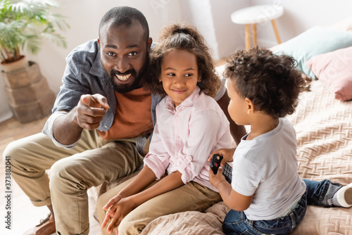 excited african american man pointing with finger near children sitting on bed