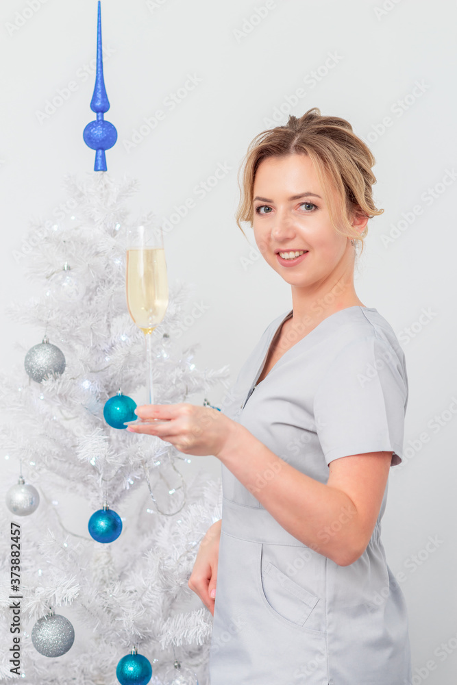 Beautiful young caucasian smiling female doctor in protective gloves with glass of champagne standing near the christmas tree Beautiful young caucasian smiling female doctor in protective gloves with glass of champagne standing near the christmas tree