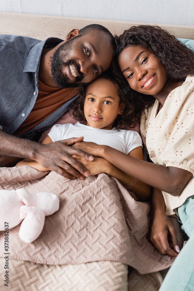 high angle view of african american husband and wife touching hands of daughter lying in bed