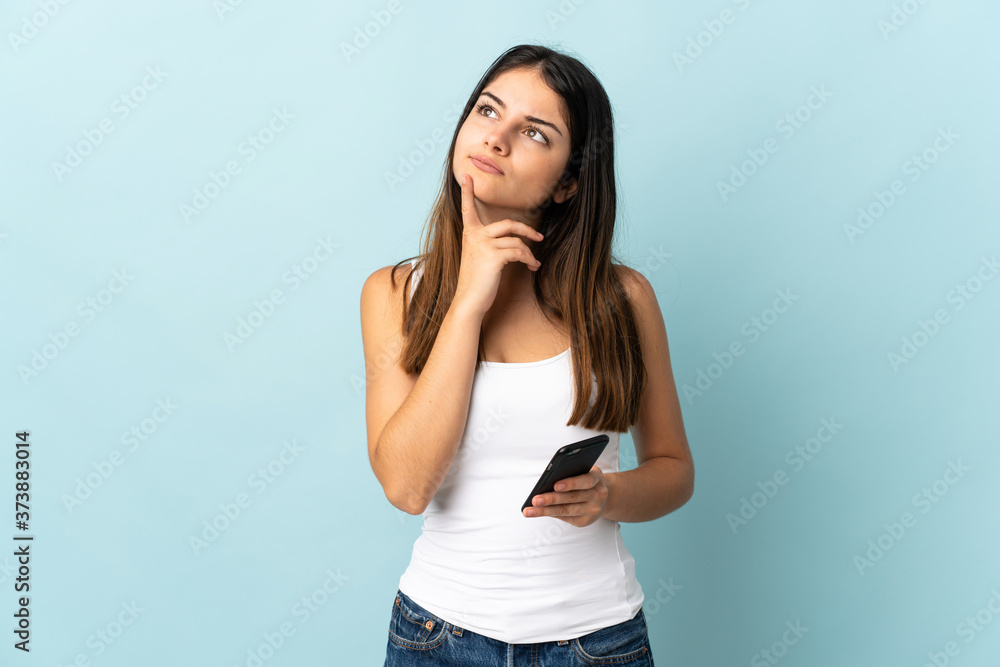 Young caucasian woman using mobile phone isolated on blue background having doubts while looking up Young caucasian woman using mobile phone isolated on blue background having doubts while looking up