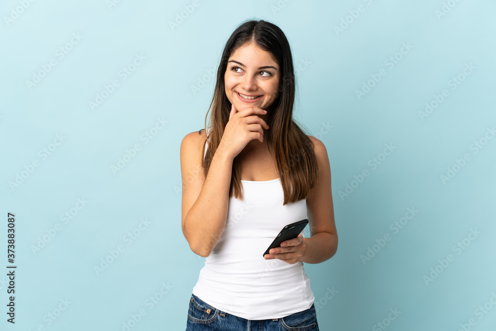 Young caucasian woman using mobile phone isolated on blue background looking up while smiling Young caucasian woman using mobile phone isolated on blue background looking up while smiling