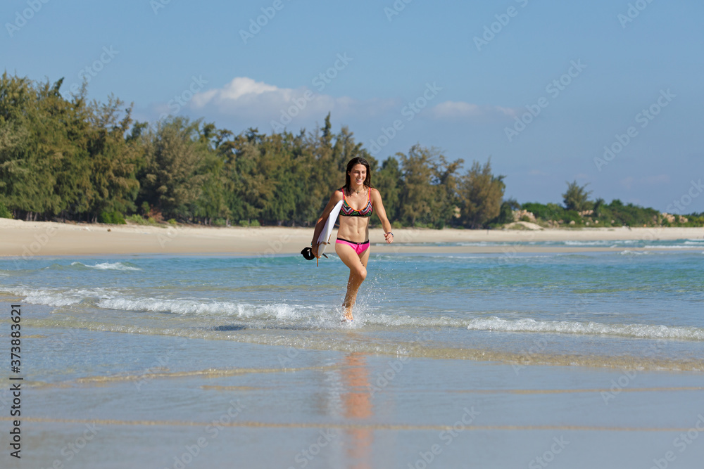 Fototapeta premium young slender and tanned girl in a swimsuit with a surfboard in her hands on the beach near the water, sea, summer, heat, sunny day, clear sea water, wave, lifestyle, sports, leisure, vacation, weeken