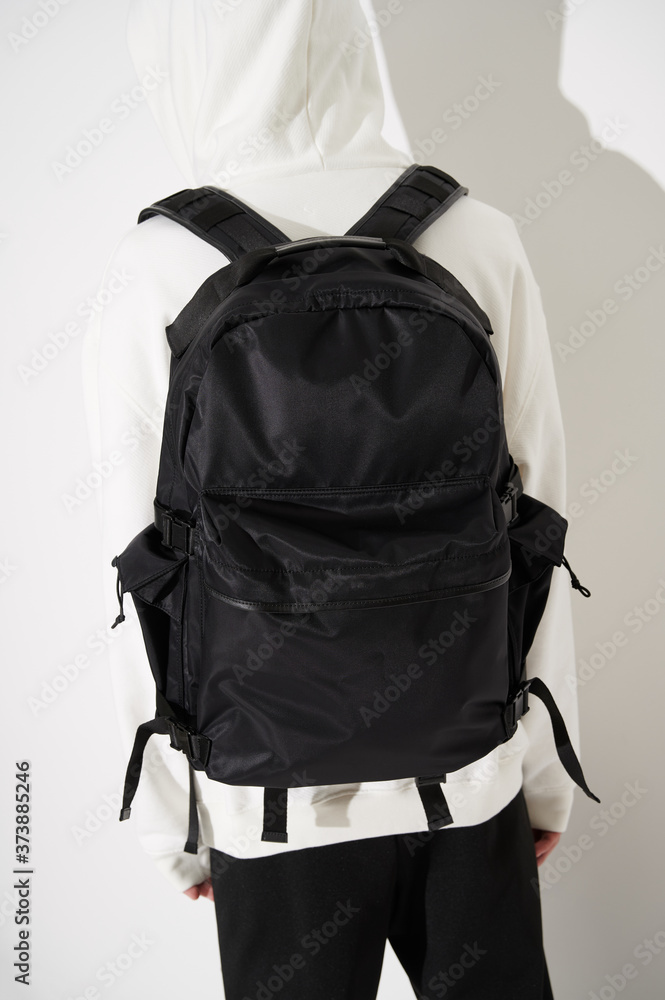 Unknown man in white hoodie with hood on and stylish black backpack near the white wall. Hard light with shadow. Fashion background. Back view. Unknown man in white hoodie with hood on and stylish black backpack near the white wall. Hard light with shadow. Fashion background. Back view.