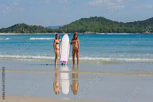 two girls in bekin with a white surfboard are standing near the sea, summer, heat, sunny day, clear sea water, wave photo