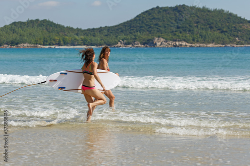 two girls in bekin with a white surfboard enter the blue sea, summer, heat, sunny day, clear sea water, wave