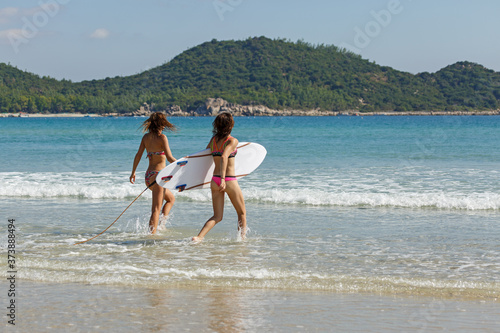 two girls in bekin with a white surfboard enter the blue sea, summer, heat, sunny day, clear sea water, wave photo