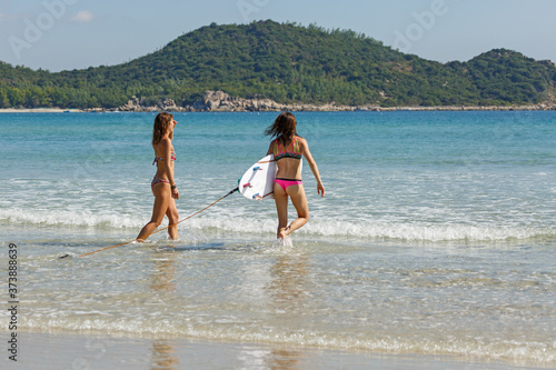 two girls in bekin with a white surfboard enter the blue sea, summer, heat, sunny day, clear sea water, wave