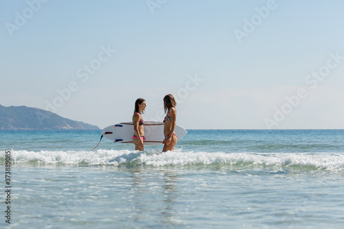 two girls in bekin with a white surfboard enter the blue sea, summer, heat, sunny day, clear sea water, wave