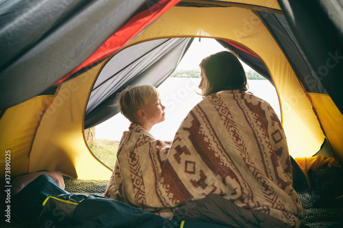 Mother and son sitting in camping tent, wrapped in wool blanket and admiring sunrise on the river. Family weekend outdoor, local travel on nature, trekking, camp lifestyle.