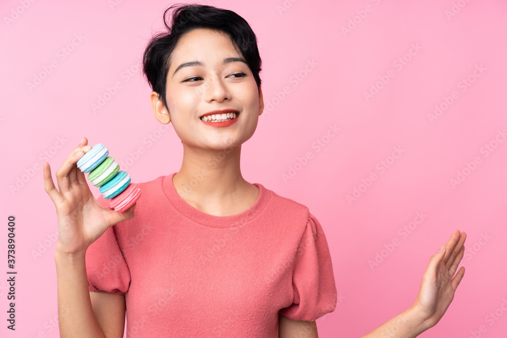 Young Asian girl over isolated pink background holding colorful French macarons and saluting Young Asian girl over isolated pink background holding colorful French macarons and saluting