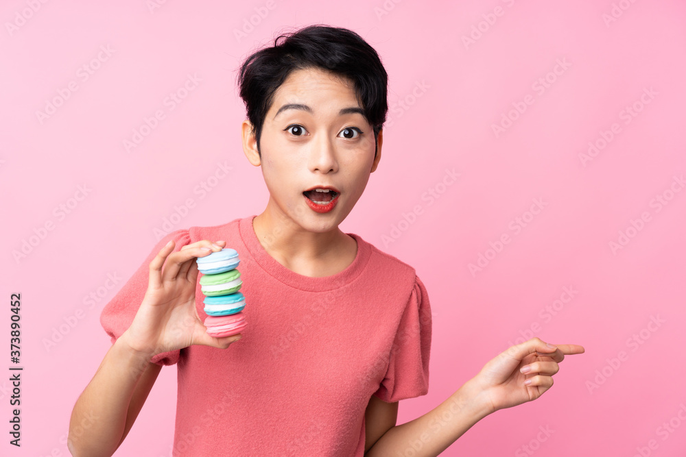 Young Asian girl over isolated pink background holding colorful French macarons and pointing side Young Asian girl over isolated pink background holding colorful French macarons and pointing side