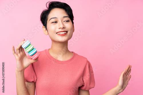 Young Asian girl over isolated pink background holding colorful French macarons and saluting