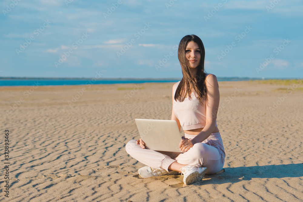 Nice woman in rose tracksuit working on a laptop on the beach.