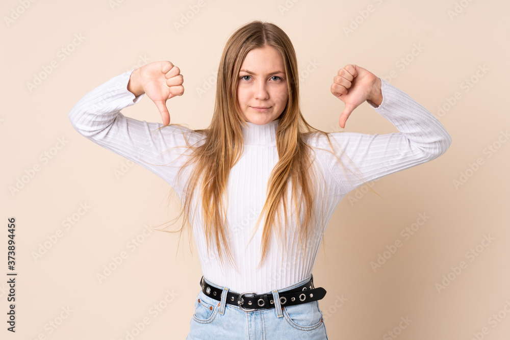 Teenager Ukrainian girl isolated on beige background showing thumb down with two hands Teenager Ukrainian girl isolated on beige background showing thumb down with two hands