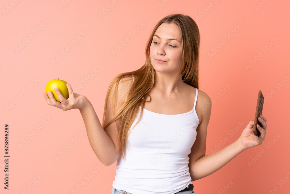 Teenager Ukrainian girl isolated on pink background having doubts while taking a chocolate tablet in one hand and an apple in the other Teenager Ukrainian girl isolated on pink background having doubts while taking a chocolate tablet in one hand and an apple in the other