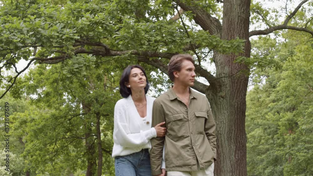 Medium shot of young Caucasian man and woman wearing casual clothes are walking holding hands in park and looking around Medium shot of young Caucasian man and woman wearing casual clothes are walking holding hands in park and looking around