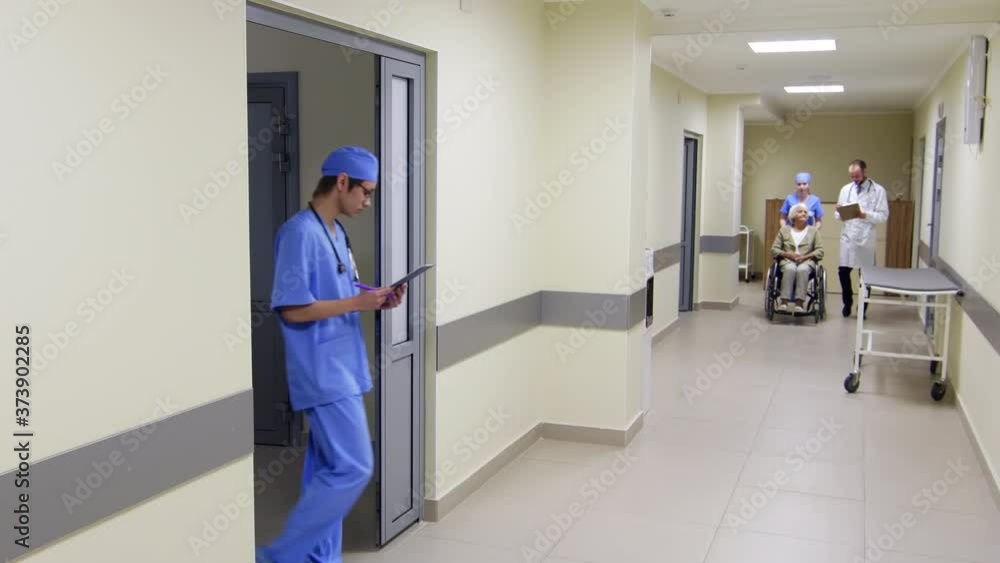 Male doctor and young nurse walking through hospital corridor with senior woman on wheelchair and discussing her treatment plan Male doctor and young nurse walking through hospital corridor with senior woman on wheelchair and discussing her treatment plan
