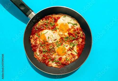 Shakshuka - poached eggs in tomato sauce, onion, pepper and spices in iron pan on blue bright background. Famous traditional Arabic and Israeli breakfast - chakchouka. Top view.
