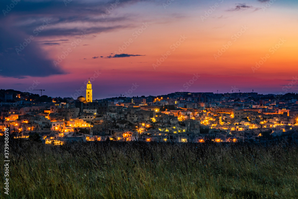 The spectacular fiery sunset and the panorama of the Sassi di Matera in Basilicata, Italy. The top view of the city from the park of the Rupestrian Churches. The lights come on as night falls.