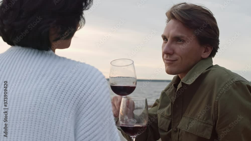 Rear view of black-haired attractive young woman and her husband are sitting on river bank in front of each other and drinking wine Rear view of black-haired attractive young woman and her husband are sitting on river bank in front of each other and drinking wine