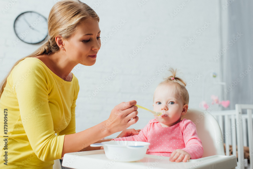 Woman holding spoon of baby food while feeding daughter on highchair at home Woman holding spoon of baby food while feeding daughter on highchair at home
