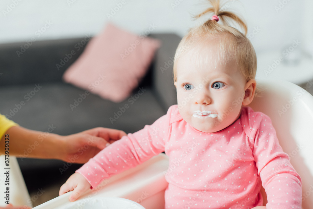 Selective focus of baby girl with messy mouth sitting on feeding chair near mother at home