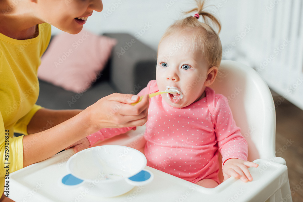 Selective focus of woman feeding baby girl on highchair at home Selective focus of woman feeding baby girl on highchair at home