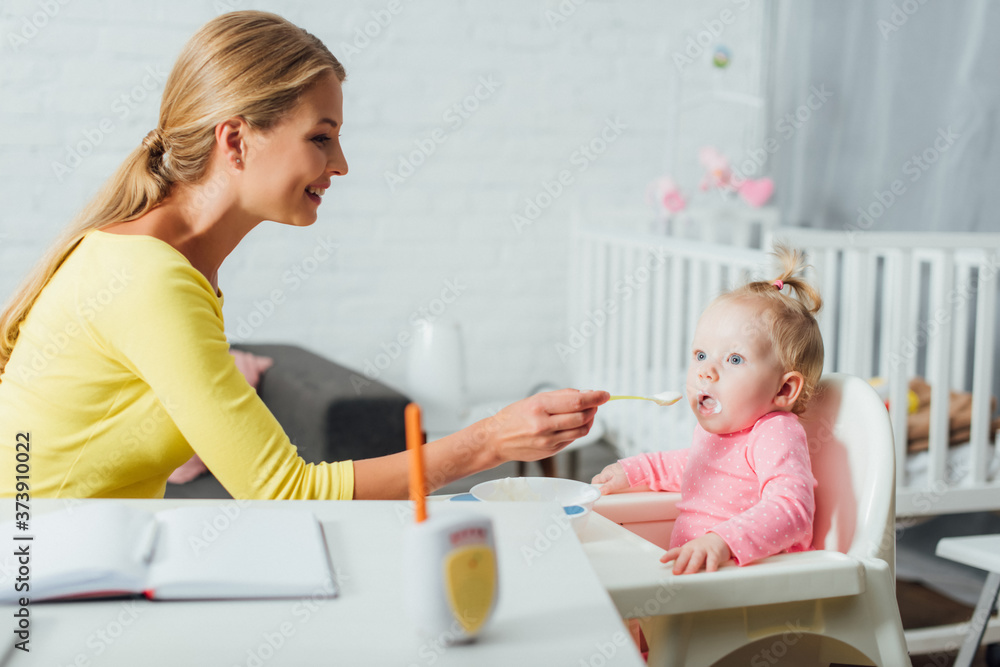 Fototapeta premium Selective focus of mother feeding baby girl on highchair near notebook on table