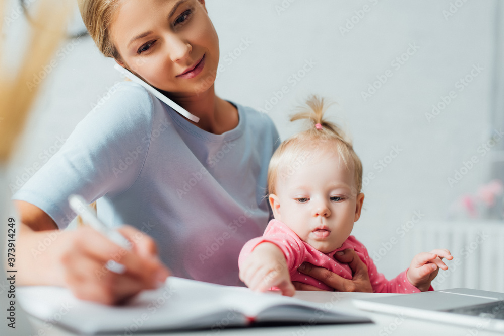 Selective focus of woman talking on cellphone while working near baby daughter at table Selective focus of woman talking on cellphone while working near baby daughter at table