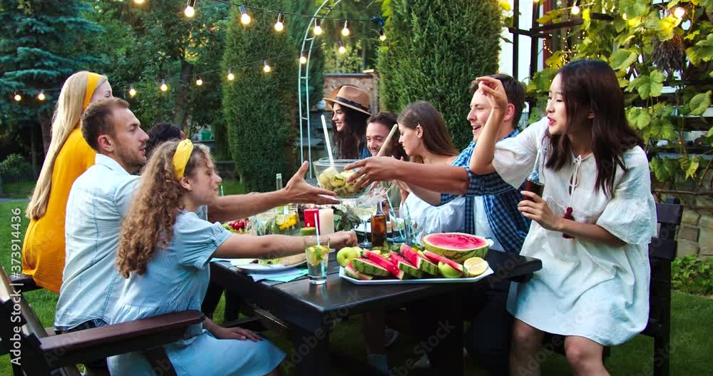 Young multi-ethnic people together eating and drinking at garden party. African American and Caucasian girls laughing. Joyful guys and Asian female chatting. Friends passing dish. Fun concept Young multi-ethnic people together eating and drinking at garden party. African American and Caucasian girls laughing. Joyful guys and Asian female chatting. Friends passing dish. Fun concept