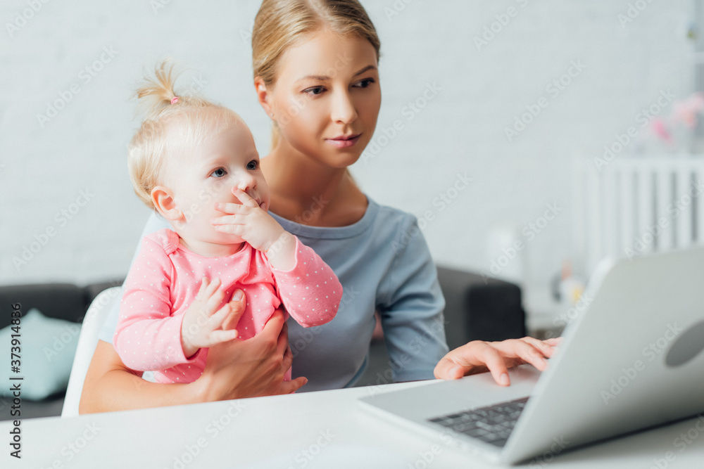 Selective focus of woman holding daughter and using laptop at table Selective focus of woman holding daughter and using laptop at table