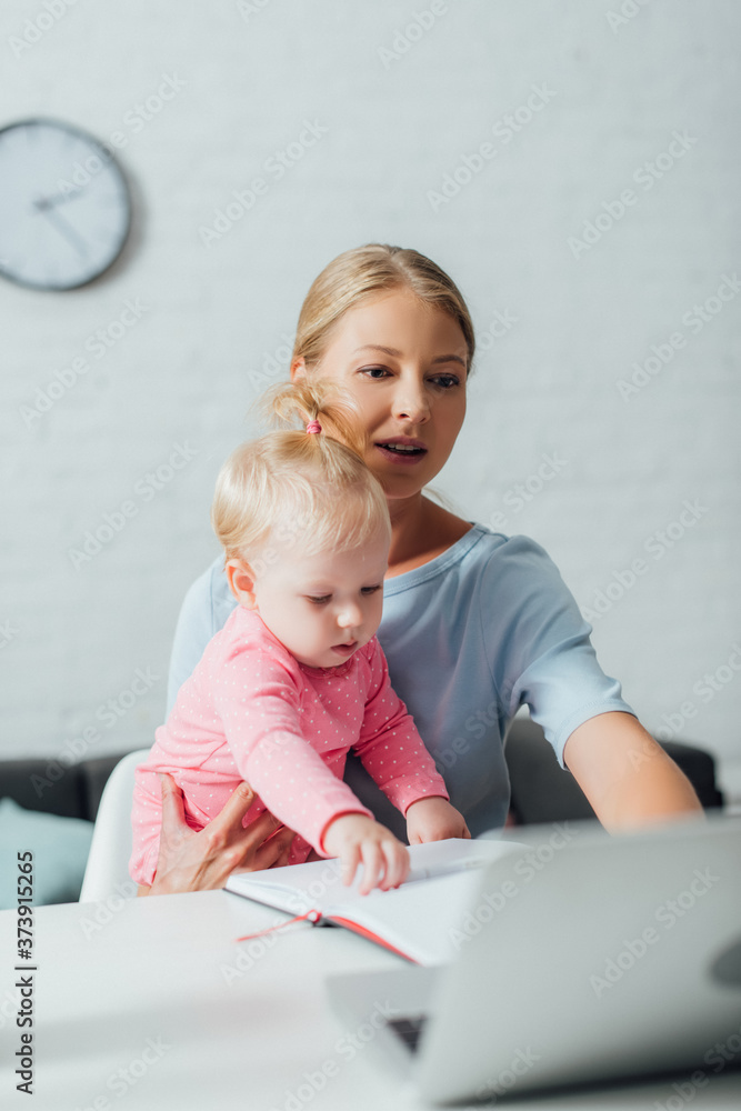 Selective focus of woman using laptop while holding baby daughter at home Selective focus of woman using laptop while holding baby daughter at home