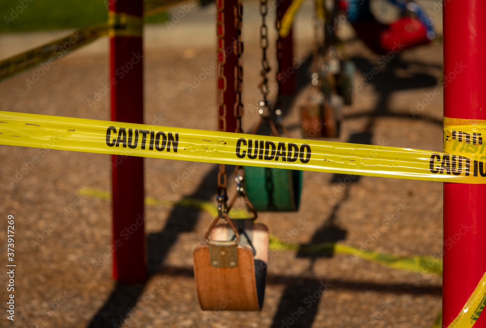 Empty saddle style swings at a playground covered in yellow caution tape written in english and spanish.