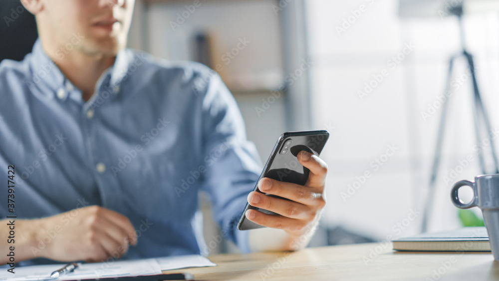 Entrepreneur Sitting at His Desk Works with Documents in the Stylish Office, Using Smartphone, Uses Social Media App, Emailing Business Partners, Messaging. Entrepreneur Sitting at His Desk Works with Documents in the Stylish Office, Using Smartphone, Uses Social Media App, Emailing Business Partners, Messaging.