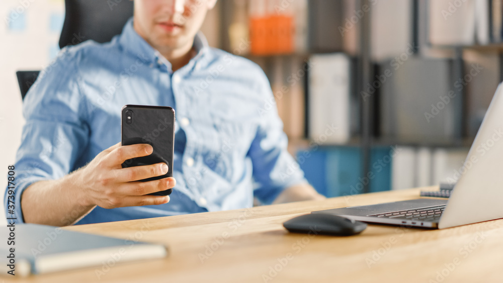 Businessman Sitting at His Desk Works on Desktop Computer in the Stylish Office and Using Smartphone, Uses Social Media App, Emailing Business Partners, Messaging. Focus On Hands and Phone Businessman Sitting at His Desk Works on Desktop Computer in the Stylish Office and Using Smartphone, Uses Social Media App, Emailing Business Partners, Messaging. Focus On Hands and Phone