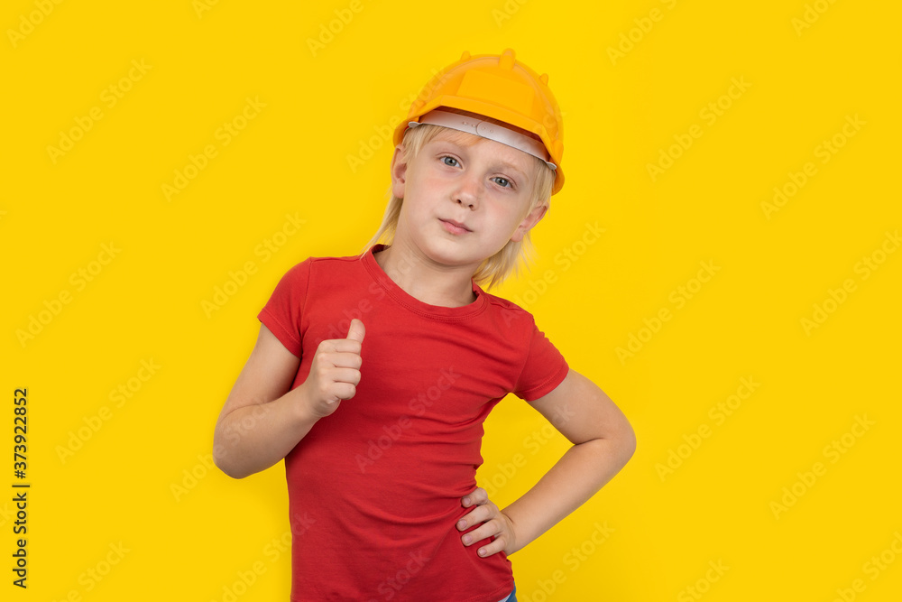 Serious teen boy in protective orange hard hat shows the gesture well done. Child on yellow background.